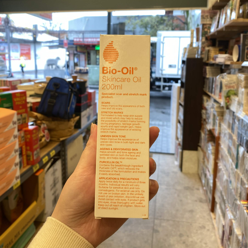 A hand holds a tall white box of Bio-Oil Skincare Oil 200ml in a retail store setting with shelves and products blurred in the background.