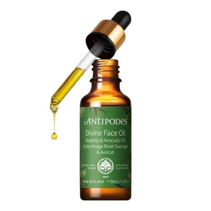 A 30ml amber glass bottle of AntiPodes Divine Face Oil with a green label, shown with a gold and black dropper cap tilted above the bottle, with a single golden oil drop hanging from the pipette against a white background.