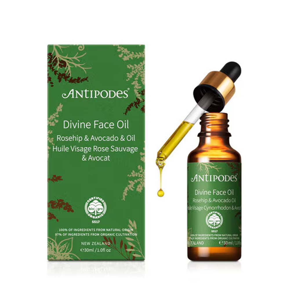 Antipodes Divine Face Oil bottle with a gold dropper and a green box, shown on a white background. The bottle has a gold neck, a green label, and the dropper is positioned above the bottle with a single drop of golden oil dripping down.